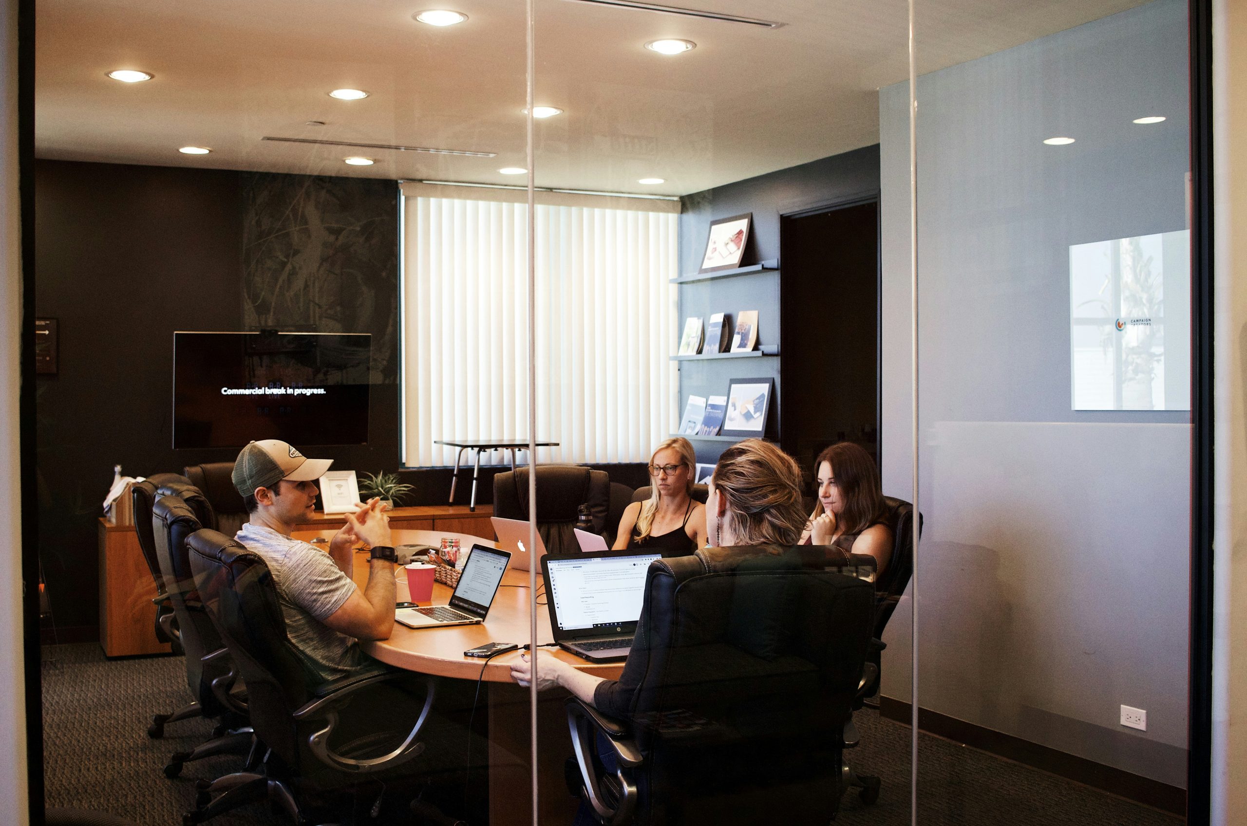 Team collaborating in a modern conference room, discussing digital marketing strategies and website design, with laptops open and a screen displaying "Commercial work in progress."