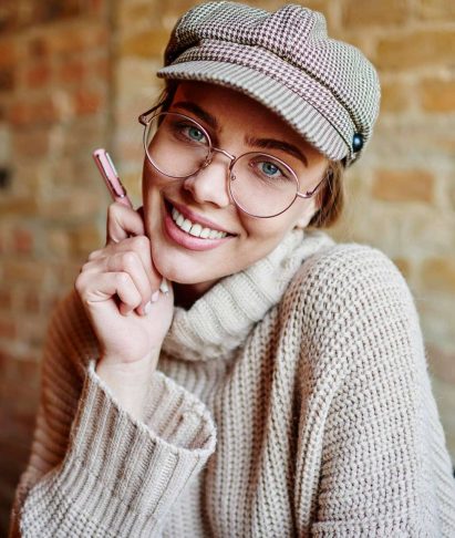 Cheerful woman in a cozy sweater and cap, smiling while holding a pen, against a brick wall background, representing the friendly and approachable team culture at Adonis Designs.